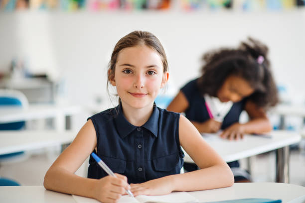 une petite fille d'école s'asseyant au bureau dans la salle de classe, regardant l'appareil-photo. - schoolgirl little girls crayon human face photos et images de collection