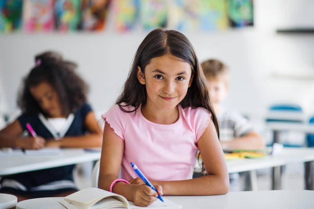 una niña pequeña de la escuela sentada en el escritorio en el aula, mirando la cámara. - schoolboy fotografías e imágenes de stock