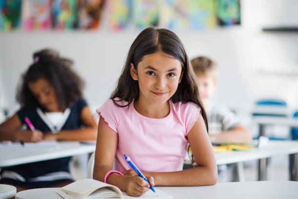 A small school girl sitting at the desk in classroom, looking at camera. A portrait of small happy school girl sitting at the desk in classroom, looking at camera. elementary school photos stock pictures, royalty-free photos & images
