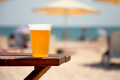 Wet glass of golden cool tasty beer on the wooden table on the shore of the sea or ocean on the sunset. Concept of beach bar or party. Alcohol beverage