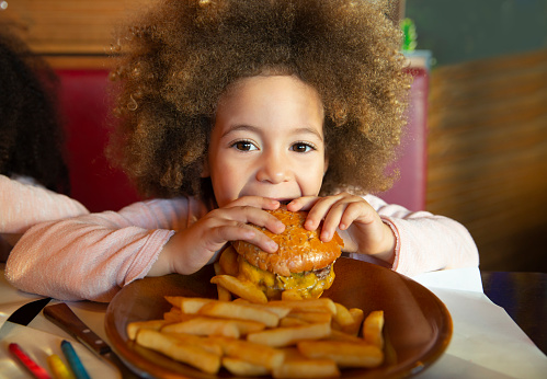 Ethnic kid girl eating burger and chips hungry at indoor restaurant