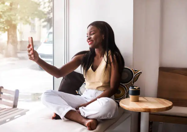 Photo of Attractive african girl makes selfie at the windowsill at cafe.