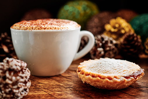 Cappuccino Coffee with a Mince Pie surrounded by pine cones and Christmas decorations on a dark rustic wood tabletop.