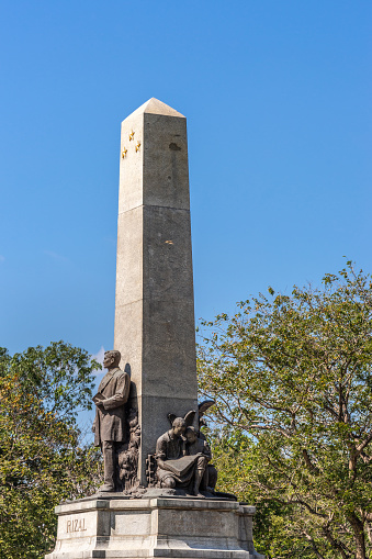 Manila, Philippines - March 5, 2019: Side closeup of Obelisk with bronze statues of Jose Rizal in Rizal Park. Green foliage in back, under blue sky.