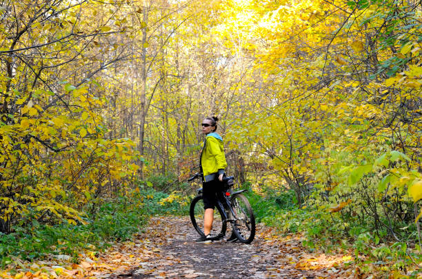 Caucasian fitness girl in a yellow jacket on a bicycle on a path in the autumn forest among yellow foliage and fallen leaves