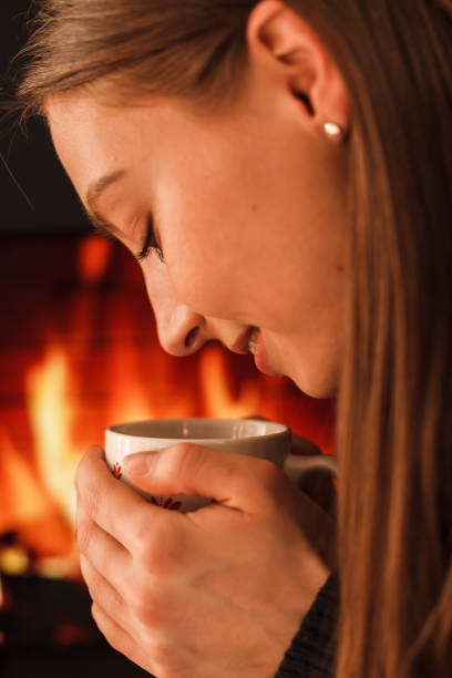 Close up of a girl holding a warm cup with both hands in front of a fireplace.