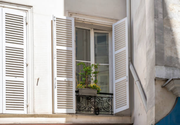 Flowers pot with flowers and plants are exposed outside a window with wooden shutters on fixed metal rims of an impromptu balcony on the facade of a plastered multilevel house