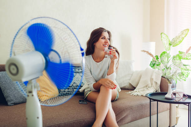 若い女性は、水を飲みながら、電話でぶら下がっている間、自宅で人工呼吸器で冷却します。夏の暑さ - summer cool electric fan heat ストックフォトと画像