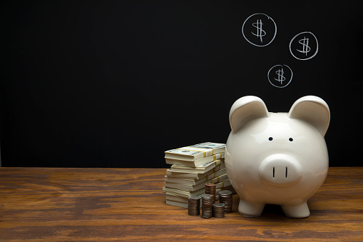 This is a photograph of a large white Piggy Bank sitting on a desk a with a male hand depositing a quarter on Black Chalkboard Background. This image could relate to savings and retirement.