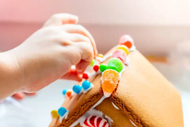 Photo of Child fingers placing candy balls and gummies on a gingerbread house roof with icing sugar as a creative and classic, seasonal holiday activity.