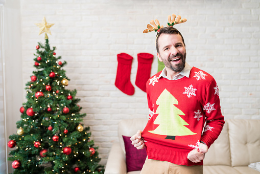 Cheerful mid adult man showing his red ugly sweaters during Christmas holiday at home