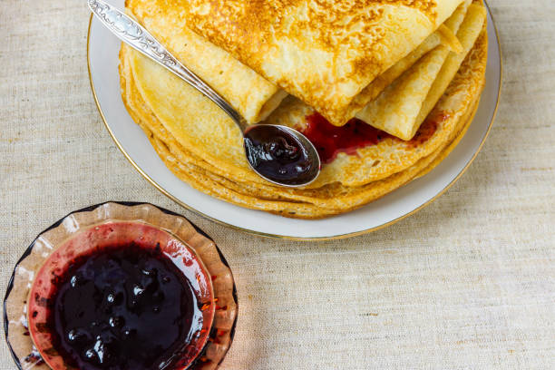 Stack of folded delicious freshly baked crepes with appetizing golden crust on white plate with homemade black current berry jam on linen table cloth