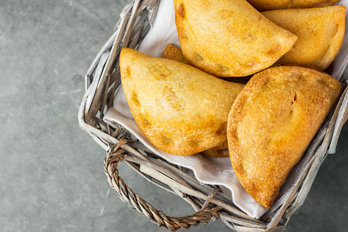 Homemade freshly baked Empanadas turnover pies with pisto vegetable cheese filling in tomato sauce in wicker basket on dark stone. Spanish Latin American cuisine savory pastry