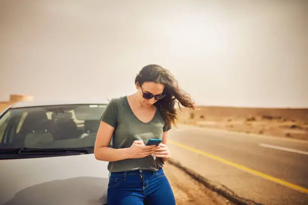 Shot of a young woman standing next to her broken down vehicle and using a smartphone