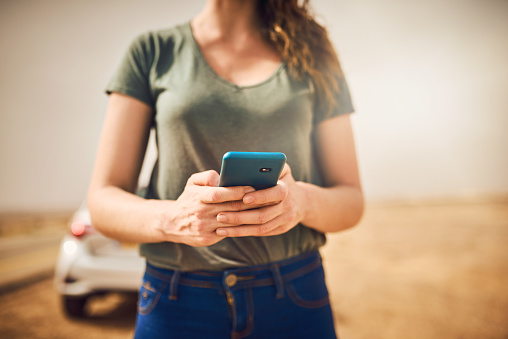 Cropped shot of a woman standing next to her broken down vehicle and using a smartphone