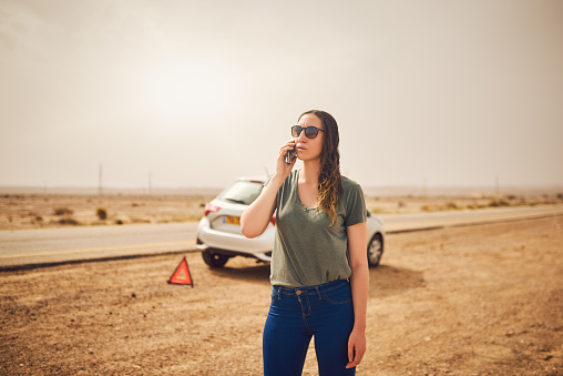 Shot of a young woman standing next to her broken down vehicle and using a smartphone