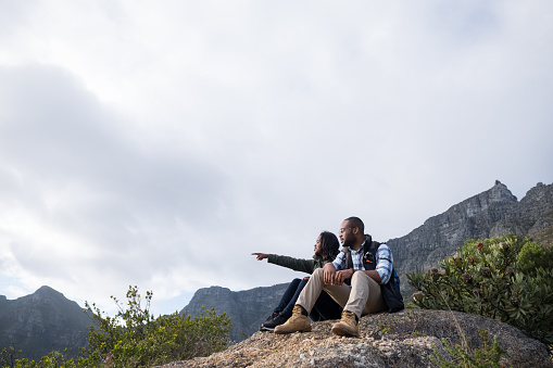 Woman gesturing while talking with boyfriend. Young couple enjoying view while sitting on rock. They on vacation at Table Mountain against sky.