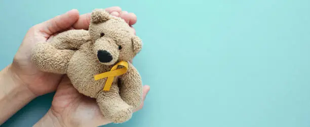 Photo of Hands holding children soft toy brown bear with yellow gold ribbon, Childhood cancer awareness