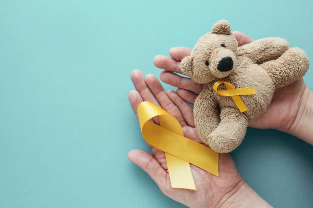 manos sosteniendo a los niños suave juguete oso marrón con cinta de oro amarillo, conciencia del cáncer de la infancia - beast cancer awareness month fotografías e imágenes de stock