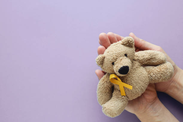 Hands holding children soft toy brown bear with yellow gold ribbon on purple background, Childhood cancer awareness stock photo