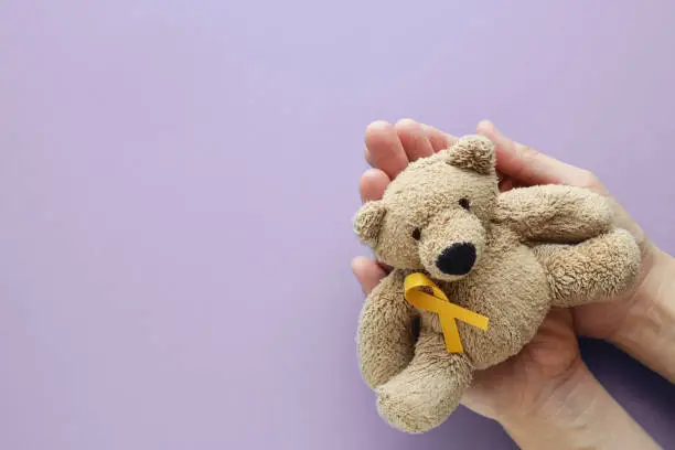 Photo of Hands holding children soft toy brown bear with yellow gold ribbon on purple background, Childhood cancer awareness