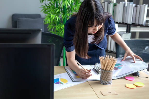 istock A beautiful Asian woman wearing a dark blue suit is taking notes in her notebook at her modern office desk and is determined to work and has a large glass window background. 1173494008