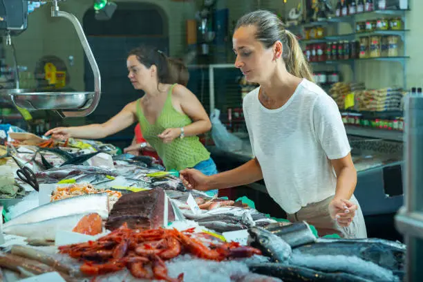 positive young female customers choosing fresh raw seafoods from large assortment of fish store