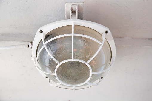 Old deck lamp with round glass shade and metal vintage protective cage on the white ceiling on the motor ship