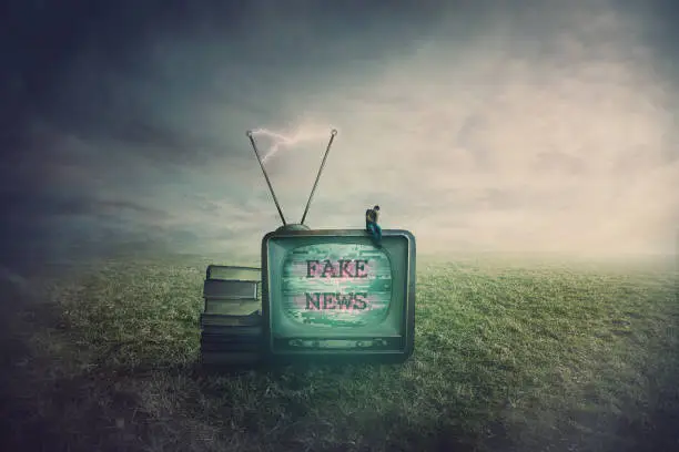Photo of Surreal scene as a minuscule man seated on an old TV box in a open field, feeling disappointed of the fake news. Television manipulation and brainwashing concept. Mass media propaganda control.