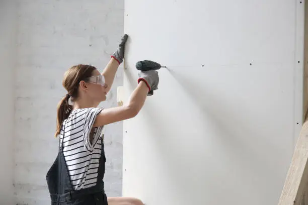 Photo of Young woman drilling screws into plasterboard with an electric screwdriver, home improvement concept