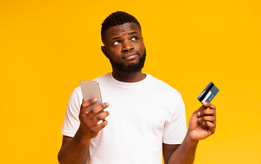 Do I longer need a credit card. Hesitant african american man holding smartphone and card, looking upwards