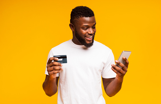 Online payment. Cheerful black man using credit card and smartphone for purchasing goods
