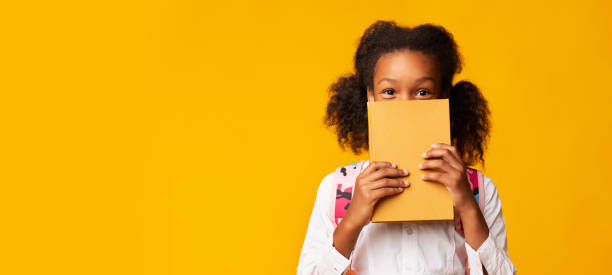 colegiala afroamericana cubriendo la cara con el libro, fondo amarillo - smart cover fotografías e imágenes de stock