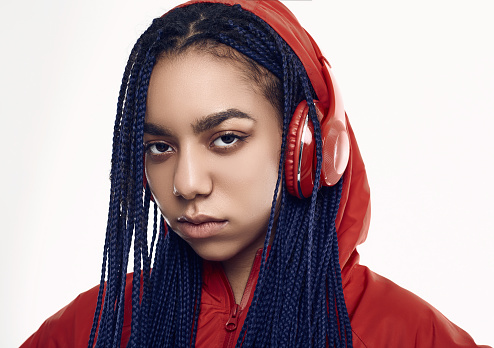 Portrait of beautiful african brunette teenage girl with dreadlocks wearing a red windbreaker listening music via wireless headphones isolated on studio background