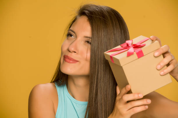 istock Young beautiful curious woman holding a present box on yellow background 1173550155