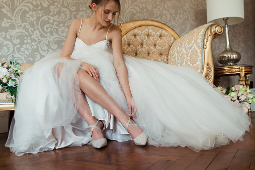 Woman dressed in her wedding gown sitting on a chair at a hotel with her skirt moved to see her shoes. She is reaching down to fasten one.