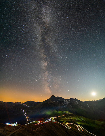light trails on curvy high alpine road through european mountains at night with moon many stars and milky way
