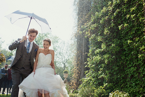Married couple laughing as they walk outside of their wedding venue as it snows. The groom is holding an umbrella above their heads. Their guests are also laughing and running along behind them.