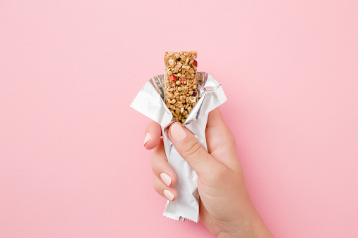 Mujer joven mano sosteniendo barra de cereales en la mesa rosa pastel. Paquete blanco abierto. Closeup. Comida dulce y saludable. Vista superior. photo