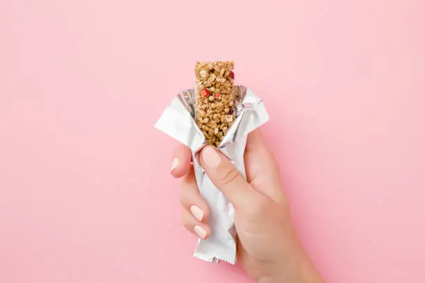 Photo of Young woman hand holding cereal bar on pastel pink table. Opened white pack. Closeup. Sweet healthy food. Top view.