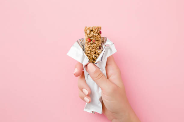young woman hand holding cereal bar on pastel pink table. opened white pack. closeup. sweet healthy food. top view. - lanchar imagens e fotografias de stock