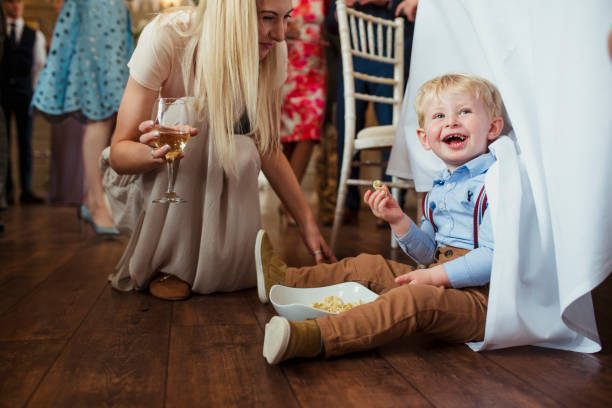 Found You! Young boy sitting under a table at a wedding reception with a bowl of snacks laughing. His mother is couching down beside him with a glass of wine in hand and smiling. family eating potato chips stock pictures, royalty-free photos & images
