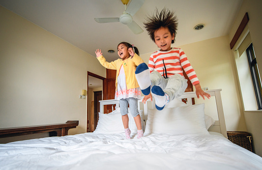 Cheerful young Chinese brother and sister jumping and playing and making mischief on messy family bed.