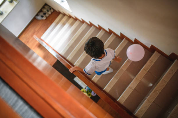 High angle rear view of young Chinese schoolboy in uniform descending staircase with pink balloon in family home.