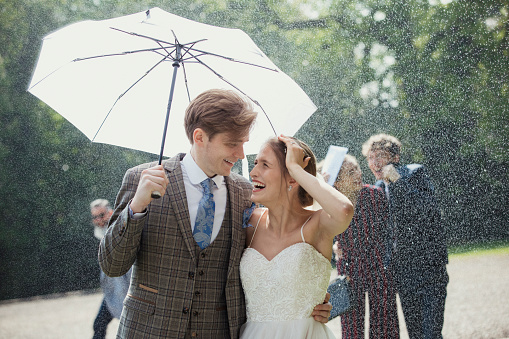 Married couple laughing and looking at each other as they walk outside of their wedding venue as it snows. The groom is holding an umbrella above their heads. Their guests are also laughing and running along behind them.