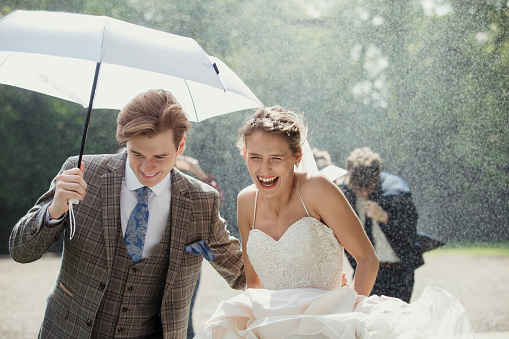 Married couple laughing as they run outside of their wedding venue as it snows. The groom is holding an umbrella above their heads. Their guests are also running along behind them.