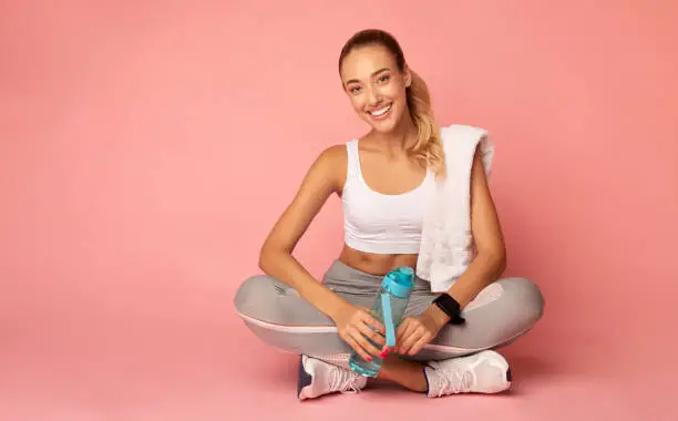 Recovery After Workout Training. Fitness Girl Resting On Floor Holding Water Bottle And Towel On Pink Background. Copy Space