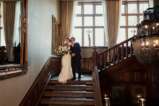 Bride linking arms with her father as they walk down the stairs on her wedding day. He is leaning over to kiss her cheek as she smiles, holding a bouquet of flowers.