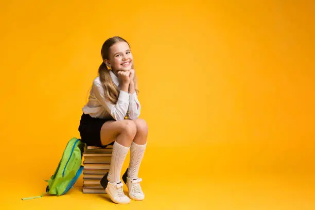 Photo of Cute School Girl Sitting On Book Stack On Yellow Background