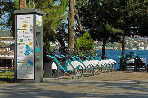 Kurucesme, Istanbul / Turkey - September 8 2019: Isbike, rent a bike service, bicycles on a bicycle stand in a public point. Sea and trees in the background. A banner with How to rent instructions on the left.